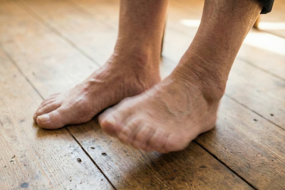 Nærbillede af en persons bare fødder, med den ene fod hævet på bolden og den anden fod lidt bagved, stående på et trægulv indendørs.