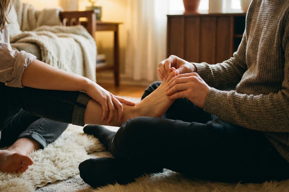 En person masserer blidt en anden persons bare fod, mens vedkommende sidder på en brun læderstol, hvilket skaber en afslappet og intim atmosfære.