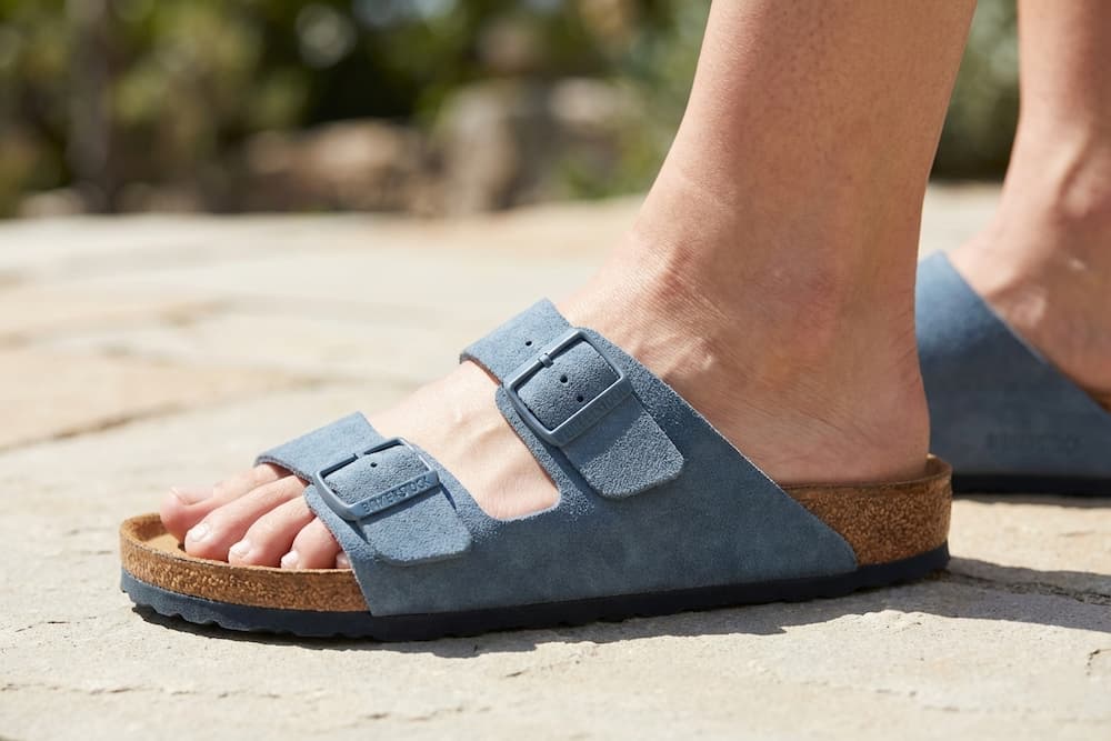 Et nærbillede af en fod iført en blå ruskindssandal med to brede remme og justerbare spænder. Sandalen har en ergonomisk korksål og vises på en lys stenbelægning udendørs, hvilket illustrerer en klassisk og komfortabel sommerstil.