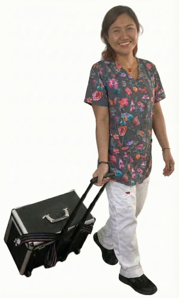 En smilende kvinde iført en grå kittel med farverigt blomstermønster og hvide bukser, der trækker en sort udstyrskuffert til fodpleje efter sig