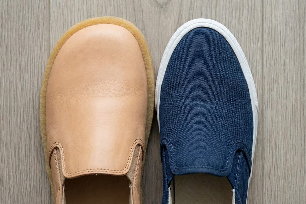 Forfra set en orange slip-on lædersko ved siden af bagfra set en blå slip-on lædersko, begge med flade brune såler, mod en ensfarvet grå baggrund.