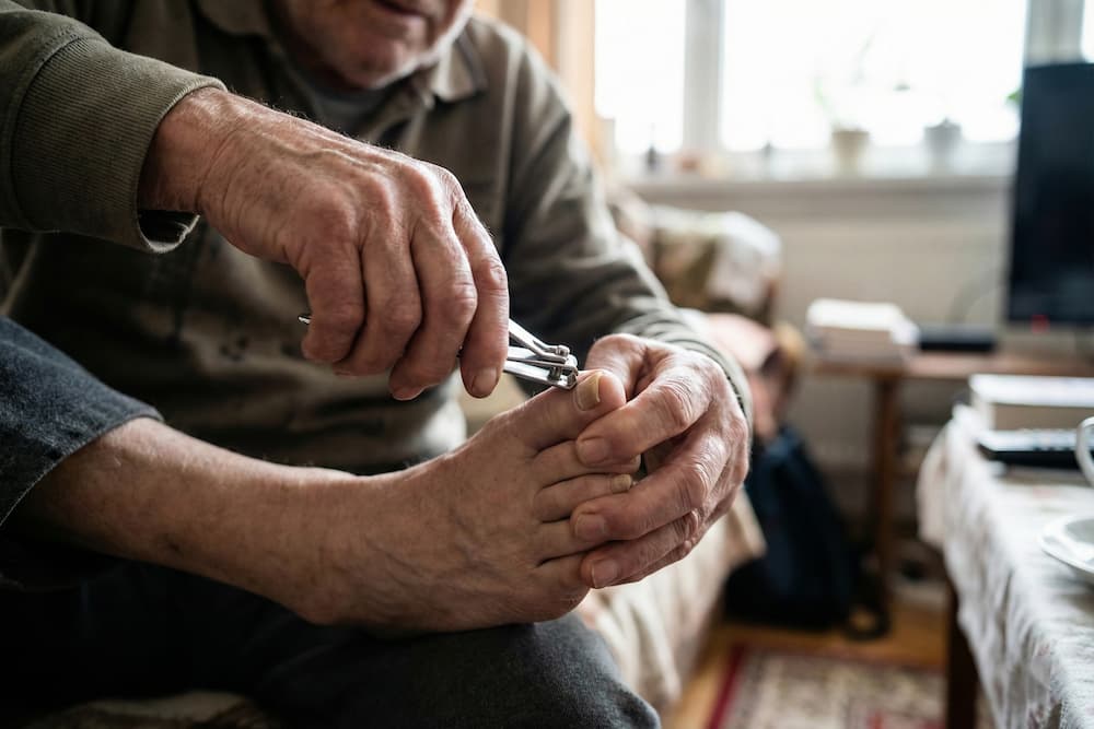 Et nærbillede af en ældre mand, der sidder foroverbøjet i en stue og kæmper med at klippe sine tånegle med en negleklipper. Hans hænder holder fast om foden, og kropsholdningen illustrerer den nedsatte mobilitet og de fysiske udfordringer, der ofte gør fodpleje vanskeligt for ældre.