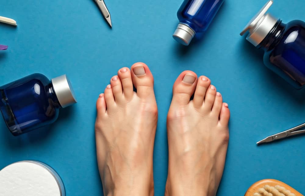 Et par bare fødder på en blå baggrund omgivet af blå flasker, negleplejeredskaber og plejetilbehør, der antyder en fodpleje- eller pedicure-setting.