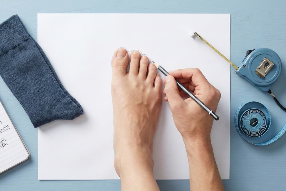 Et instruktivt billede set oppefra, der viser metoden til at måle fodstørrelse. En hånd tegner omridset af en bar fod på et hvidt ark papir med en blyant, mens en blå sok og målebånd ligger klar på den lyseblå overflade.