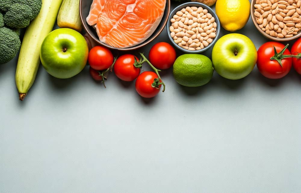 Diverse sunde fødevarer, herunder laks, hvide bønner, æbler, tomater, en agurk, en citron, en lime og broccoli, er arrangeret i en række øverst i billedet på en lyseblå baggrund.