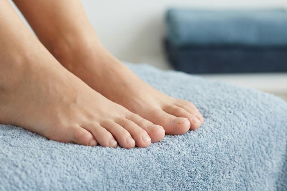 Et nærbillede af to velplejede bare fødder, der hviler på et lyseblåt frottéhåndklæde. Tåneglene er klippede, rene og glatte med en naturlig farve, hvilket illustrerer, hvordan sunde negle bidrager til fodens generelle velbefindende i et rent og hygiejnisk miljø.