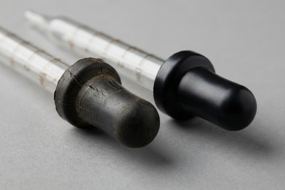 Et nærbillede, der sammenligner to glaspipetter liggende side om side. Pipetten til venstre har en mørnet, tør og revnet gummibold, mens pipetten til højre fremstår med en ny, glat og skinnende sort gummibold, hvilket tydeliggør forskellen på slidt og nyt udstyr.