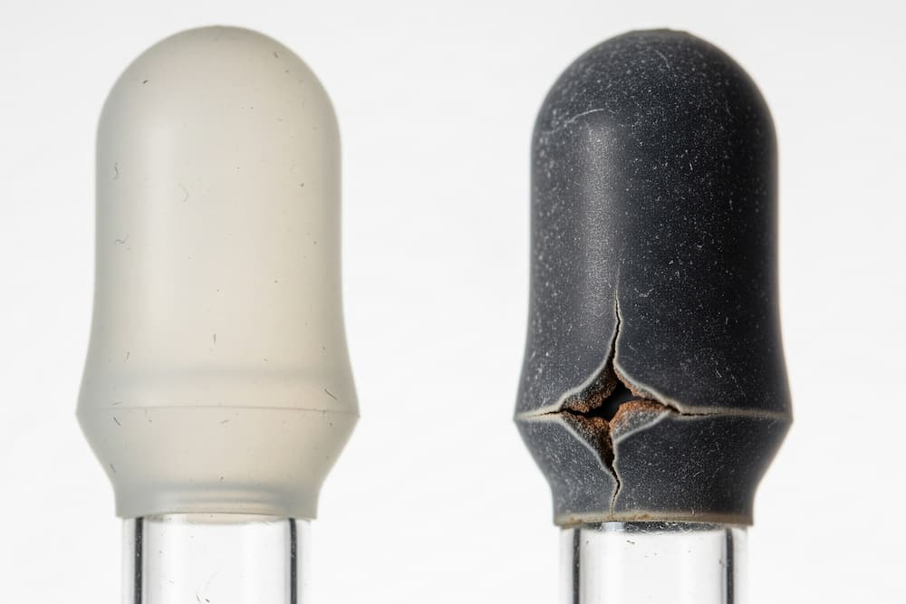 To sorte gummipærer på glasrør mod en mørk baggrund; den venstre pære er slidt og revnet med en afrundet top, mens den højre pære er nyere, glat og har en cylindrisk top.