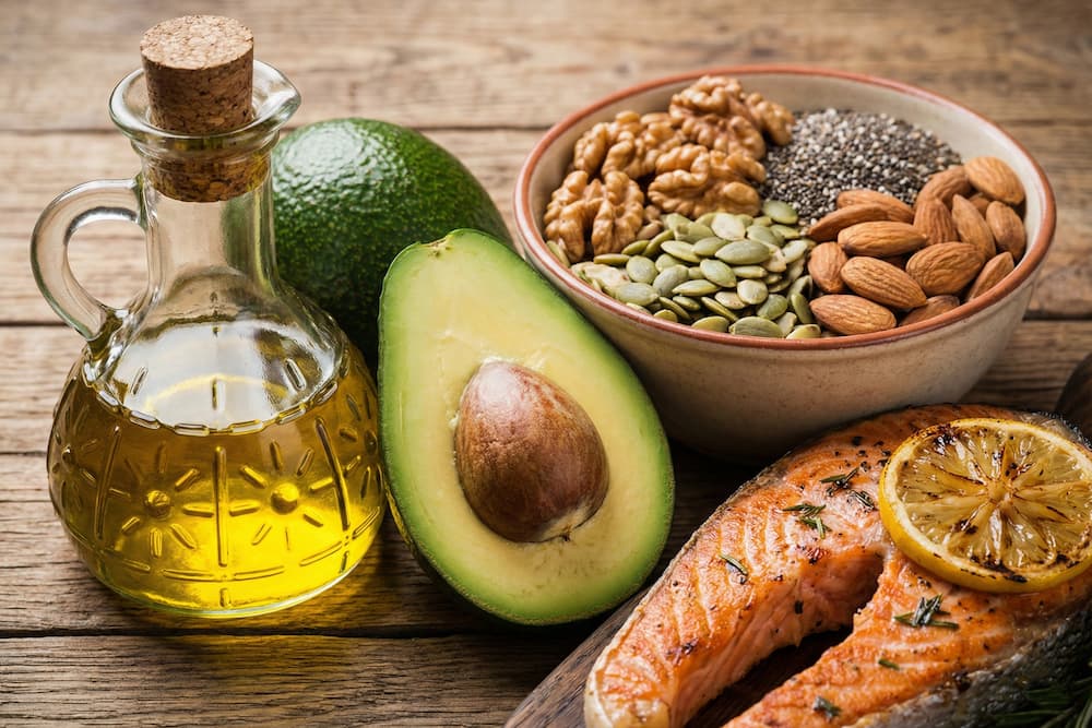 Et stilfuldt billede på en mørk baggrund, der præsenterer kilder til sunde fedtsyrer. Opstillingen inkluderer en grillet laksebøf med citron, en halv frisk avocado, en glaskaraffel med olivenolie samt en skål med blandede nødder og kerner, der er vigtige for at modvirke inflammation i kroppen.