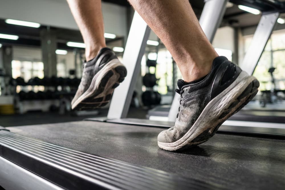 Nærbillede af en persons fødder i sorte sportssko, der går på et løbebånd, med fokus på skoene og løbebåndet i svagt oplyste indendørs omgivelser.