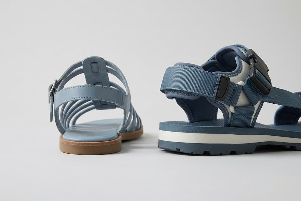 Et nærbillede, der sammenligner bagpartiet på to blå sandaler. Til venstre ses en fin lædersandal med mange remme og et spænde om anklen, mens sandalen til højre er en sporty model med en tyk, hvid sål og en bred, polstret hælrem i tekstil, hvilket illustrerer forskellen i støtte og konstruktion.