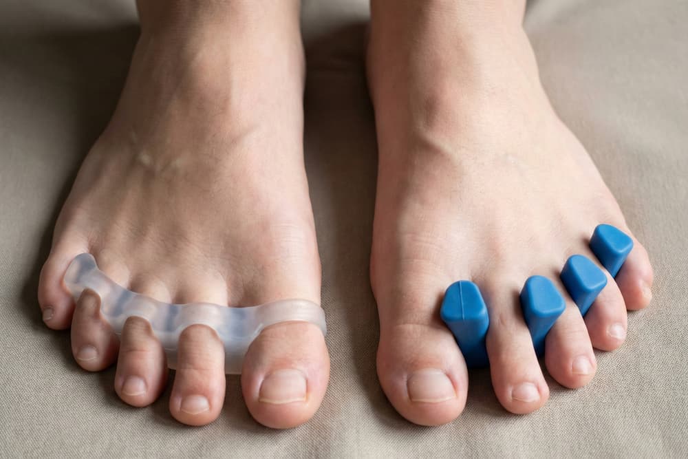 Nærbillede af to bare fødder med blå tåskillere placeret mellem tæerne, mod en neutral brun baggrund. Indretningerne er designet til at holde tæerne spredt fra hinanden.