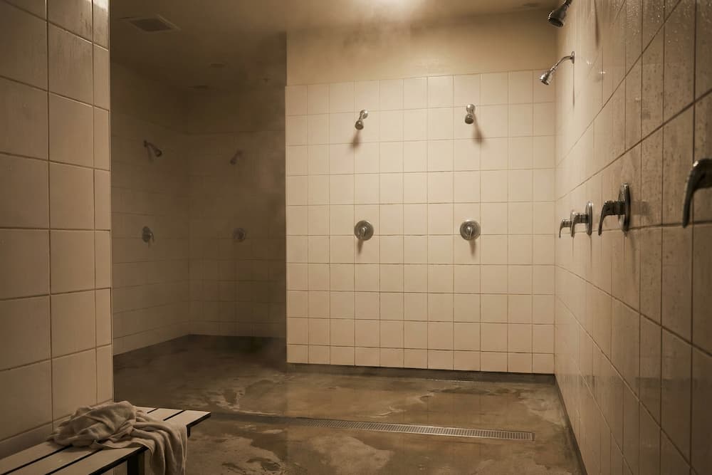 Et dunkelt og dampfyldt fællesbadeområde med hvide fliser. Gulvet er vådt med store vandpytter, og et brugt håndklæde ligger på en bænk, hvilket illustrerer det varme, fugtige miljø, hvor smitterisikoen for neglesvamp er særligt høj.